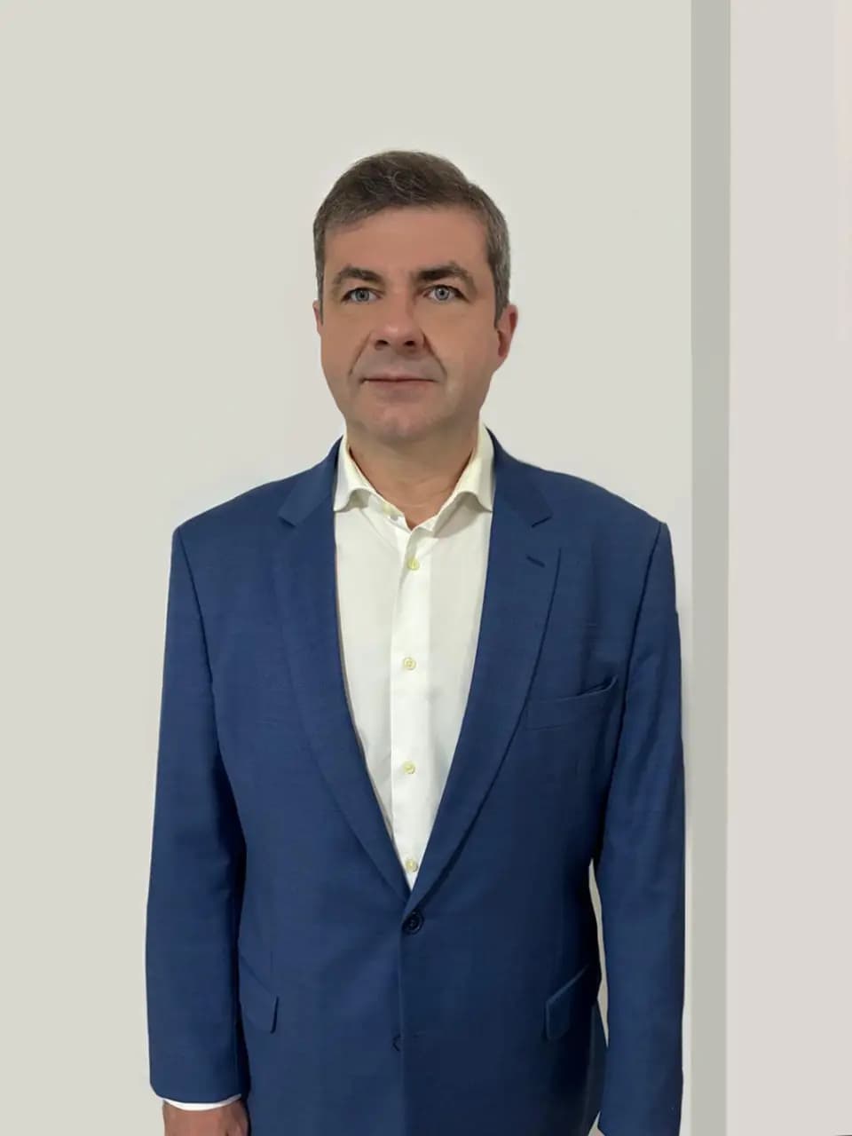Retrato vertical de Moisés Bauer, homem de pele clara, cabelos curtos castanhos com fios grisalhos e expressão serena. Ele está em pé, com o rosto levemente voltado para a esquerda, vestindo terno azul sobre camisa social branca, sem gravata. Ao fundo, há uma parede clara e neutra.